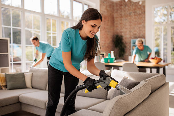 gettyimages-2214683477-612x612 Woman deep cleans a sofa using professional equipment while her colleagues clean various areas of a stylish apartment.